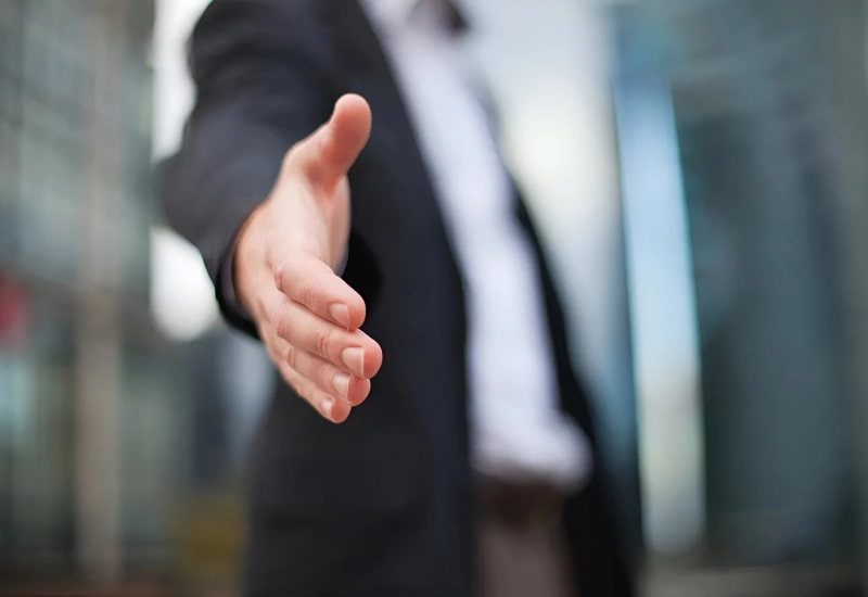 A person in a suit extending their hand forward, as if offering a handshake, signifying a commitment to revenue maximization. The background is blurred, emphasizing the importance of the hand gesture.