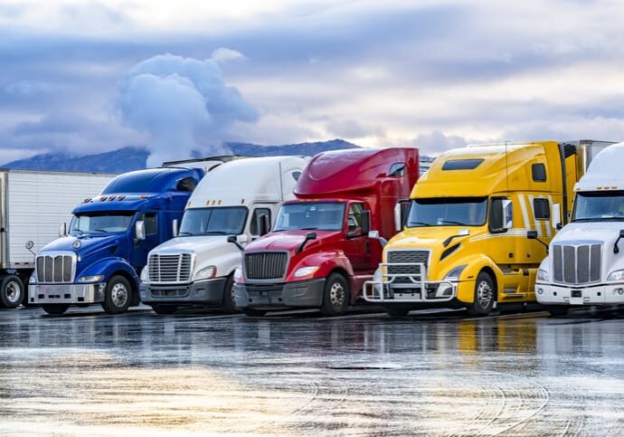 Different make big rigs semi trucks tractors with loaded semi trailers standing in the row on truck stop parking lot at early morning waiting for the route continuation time according to the log book